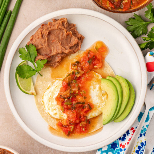 Huevos rancheros on a plate with refried beans, avocade slices, and a lime wedge.