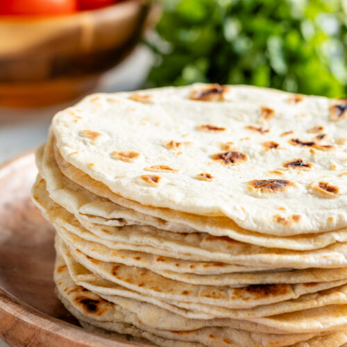 Stack of homemade flour tortillas on a plate with tomatoes and cilantro in the background.