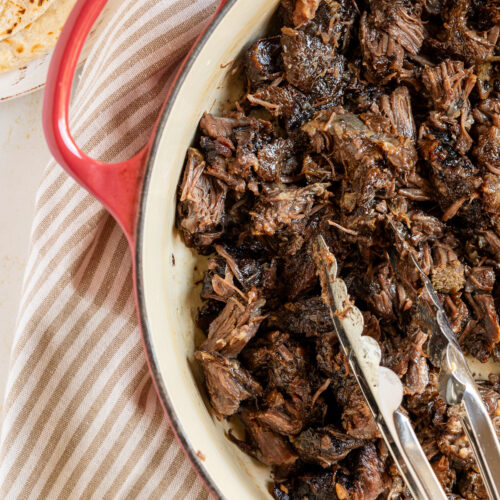 Beef barbacoa in a Dutch oven, shown with tongs.